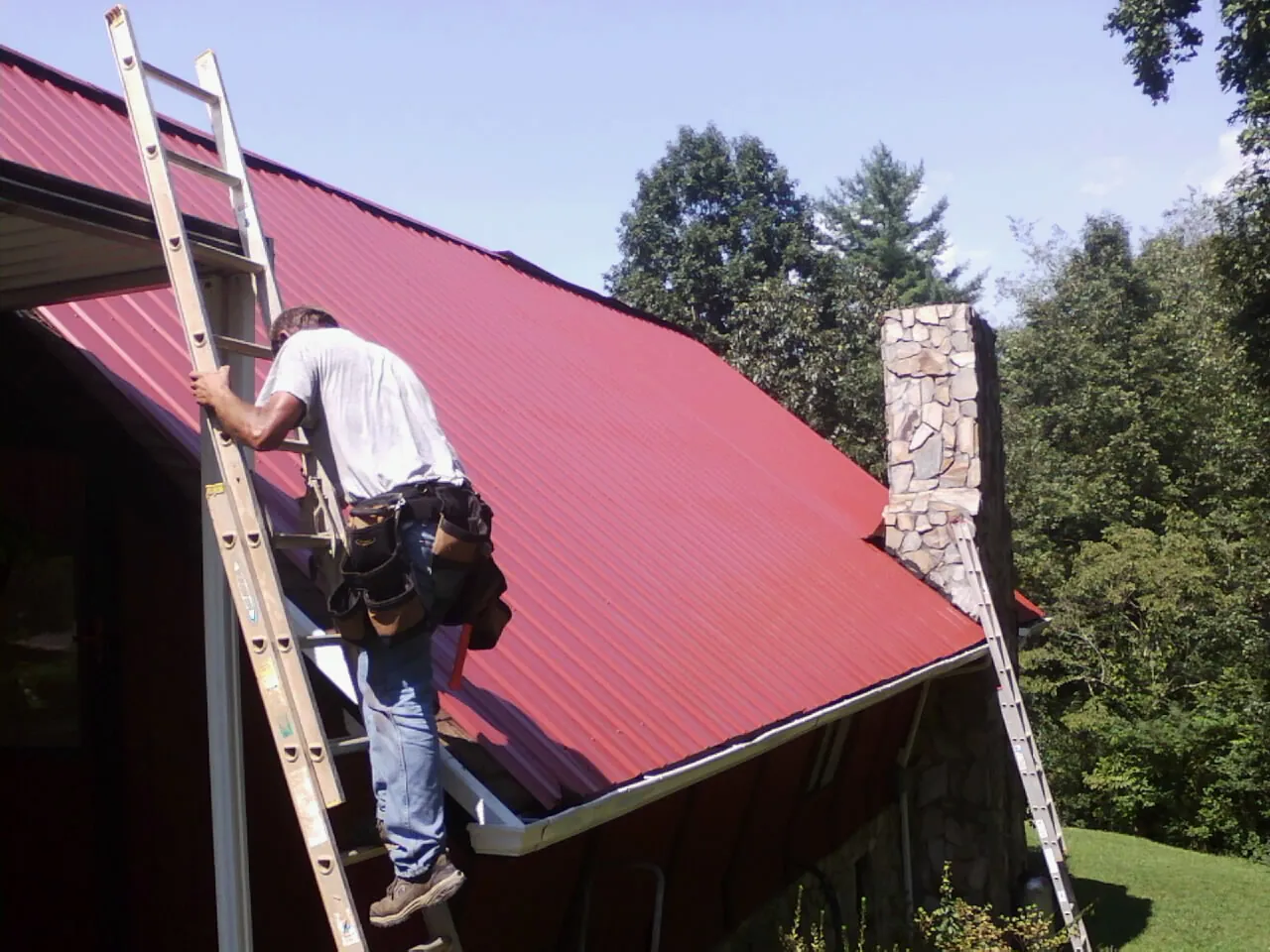 Roofer on a ladder beside a red metal roof and stone chimney — Soffit & Fascia Repair by Roofing Brothers in Chelsea
